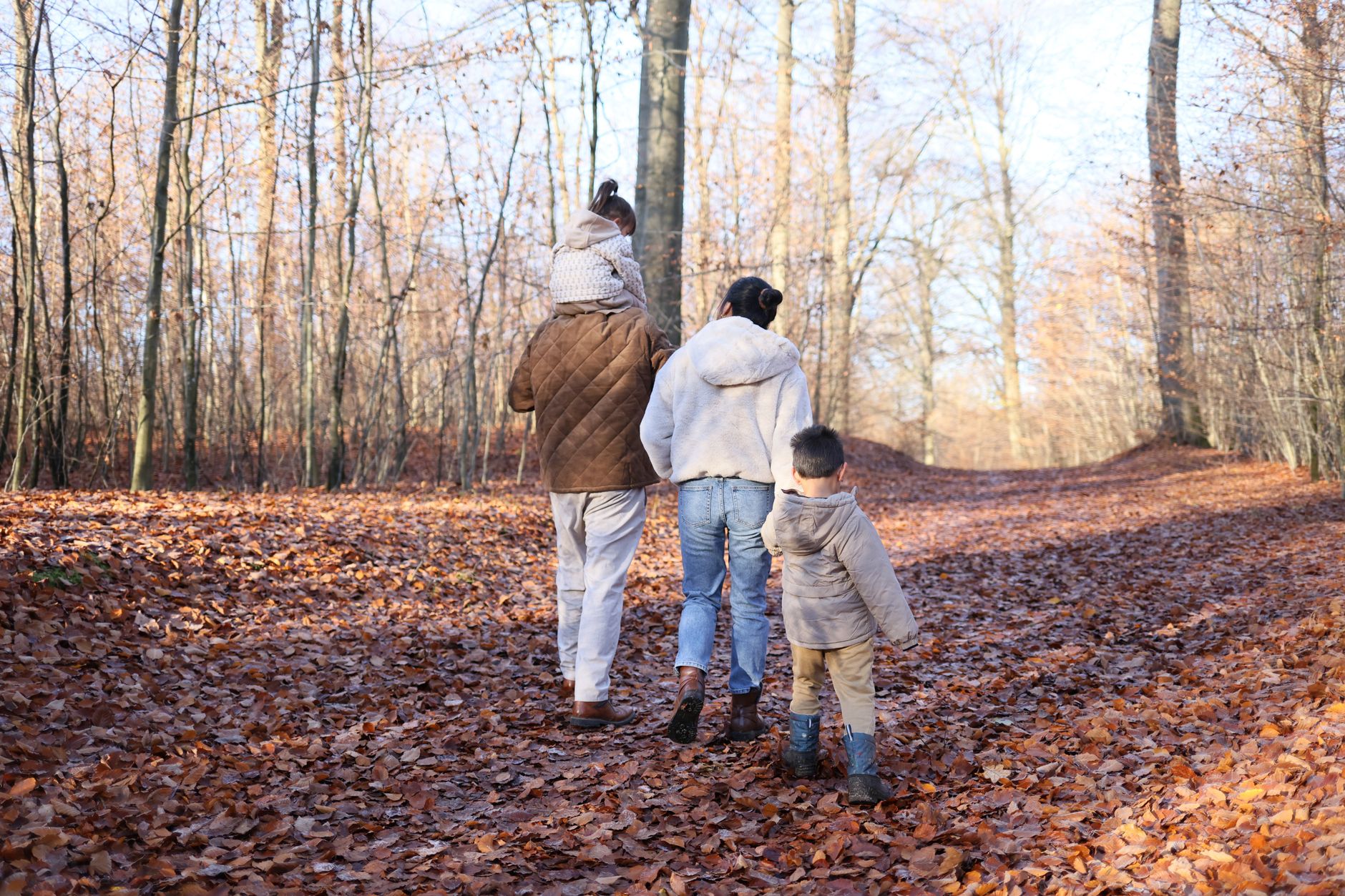Famille vue de dos marchant sur un chemin de forêt couvert de feuilles mortes, un enfant sur les épaules d’un adulte.