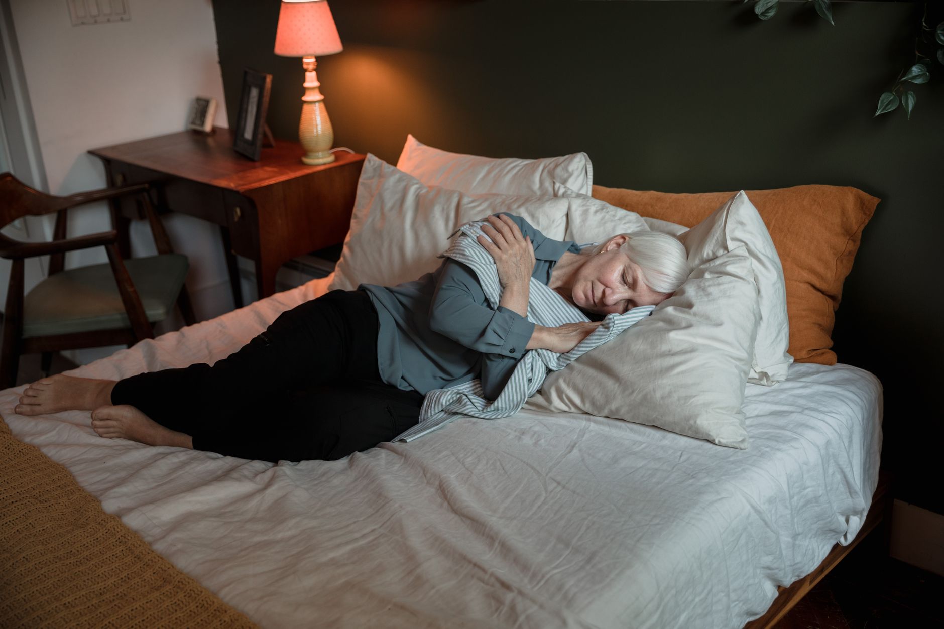 Femme âgée aux cheveux gris allongée sur un lit, les yeux fermés, entourée d’oreillers, près d’une table de chevet avec une lampe allumée.