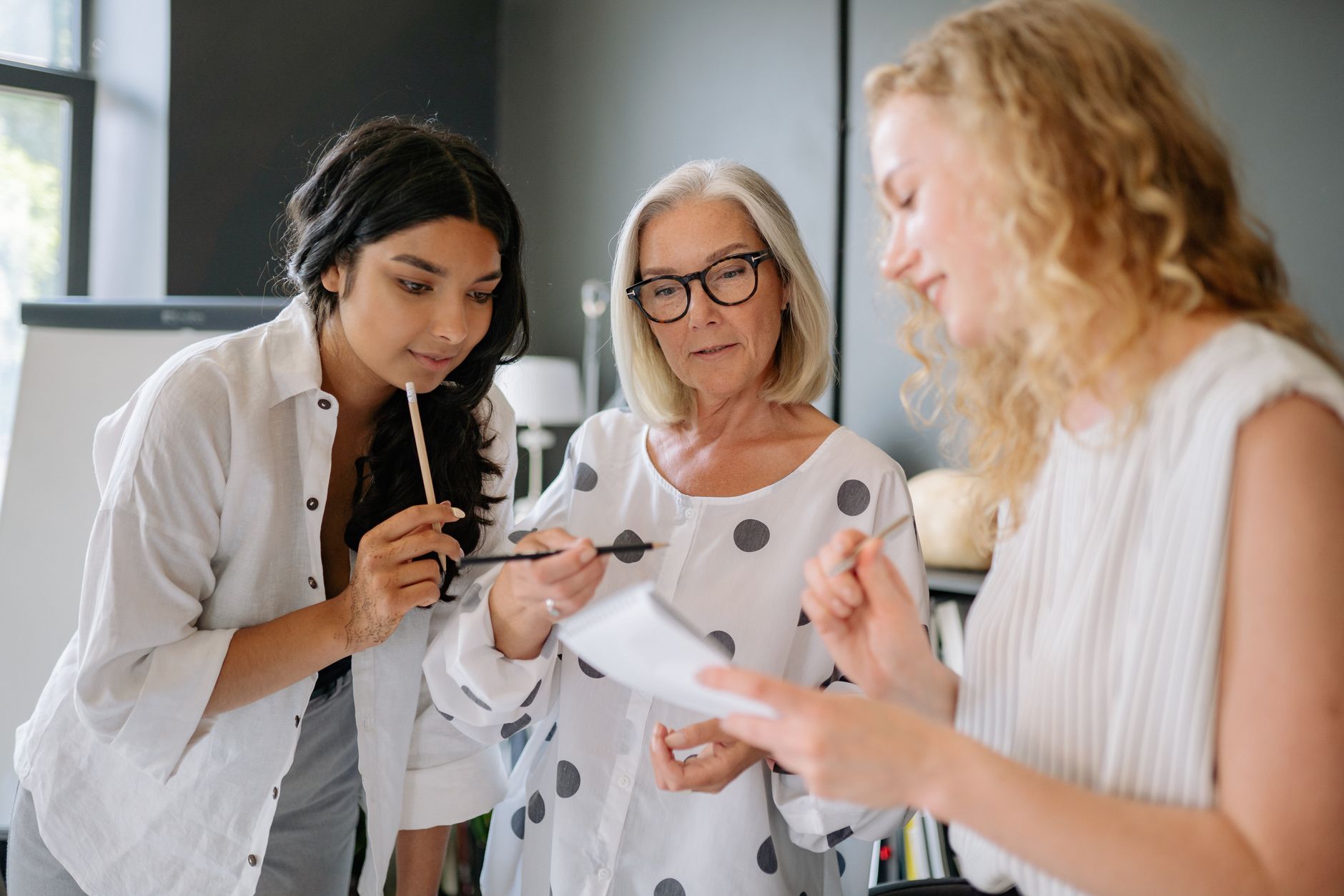 Trois femmes de différents âges se penchent sur un bloc-notes dans un bureau, un crayon à la main, lors d’un échange de travail.
