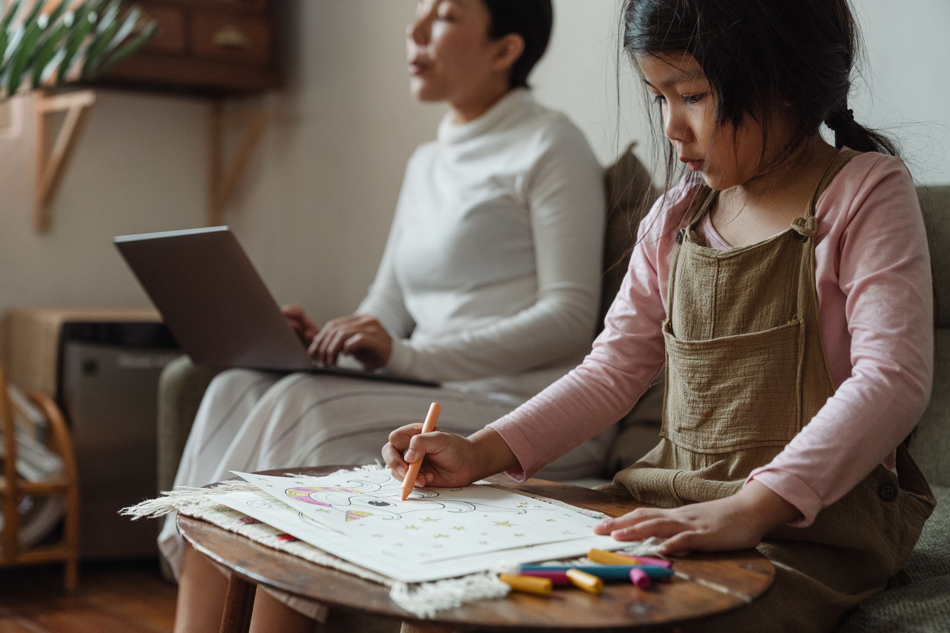 Une fillette colorie une feuille avec un crayon sur une table basse en bois, pendant qu’une femme floue tape sur un ordinateur portable derrière elle.
