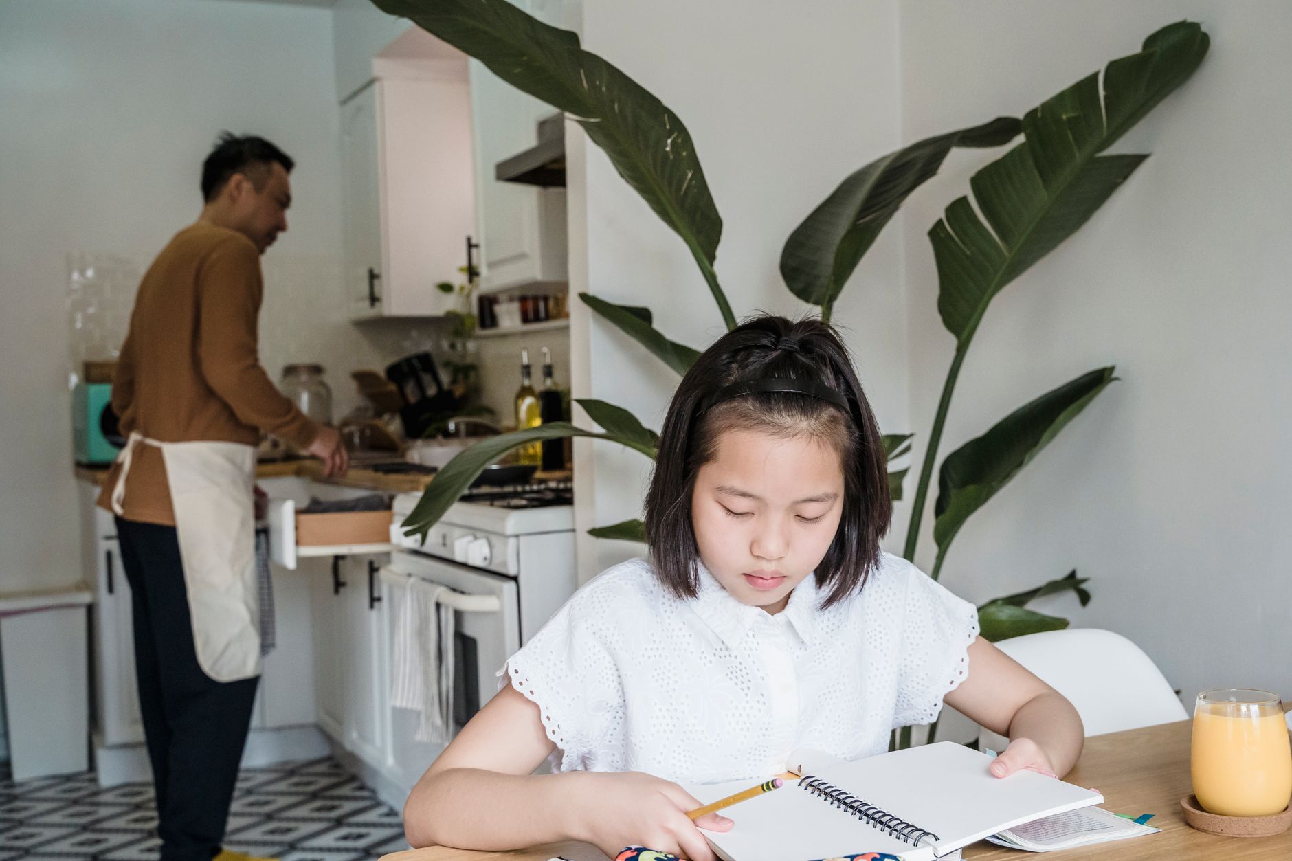 Une jeune fille fait ses devoirs avec un carnet et un crayon à table, pendant qu’un adulte prépare à manger dans la cuisine en arrière-plan.